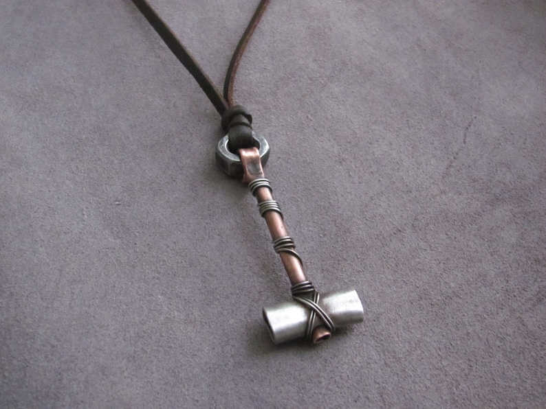 May include: A silver and copper hammer pendant necklace with a leather cord. The hammer head is made of metal and has a rounded shape. The handle is wrapped in copper wire.