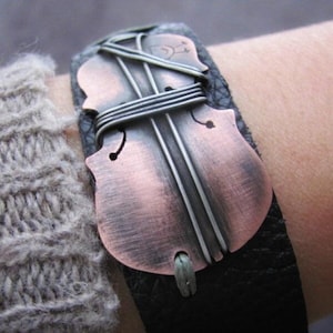 May include: A black leather bracelet with a copper and silver violin-shaped charm. The charm is wrapped with silver wire and has a distressed finish.