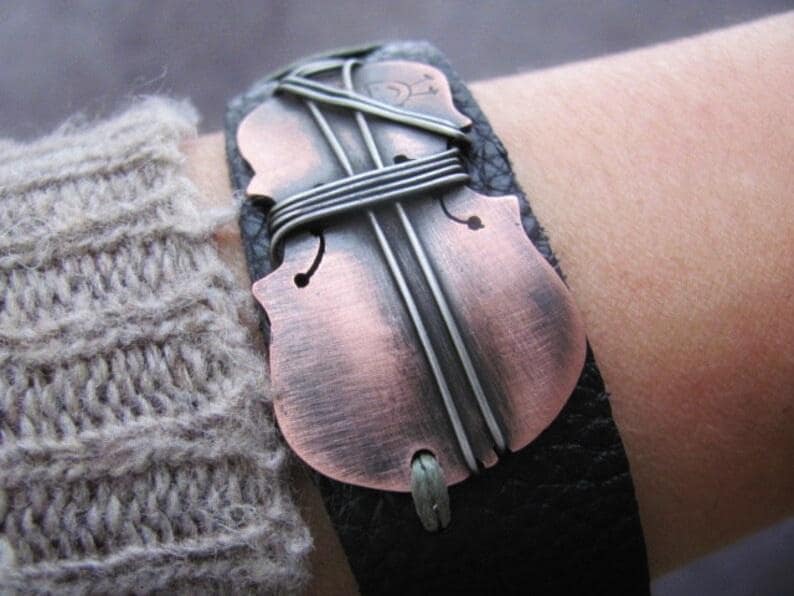 May include: A black leather bracelet with a copper and silver violin-shaped charm. The charm is wrapped with silver wire and has a distressed finish.