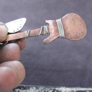 May include: A copper guitar-shaped pendant with a silver wire wrapped around the body. The pendant is attached to a silver pick with the word "I PICK" engraved on it.