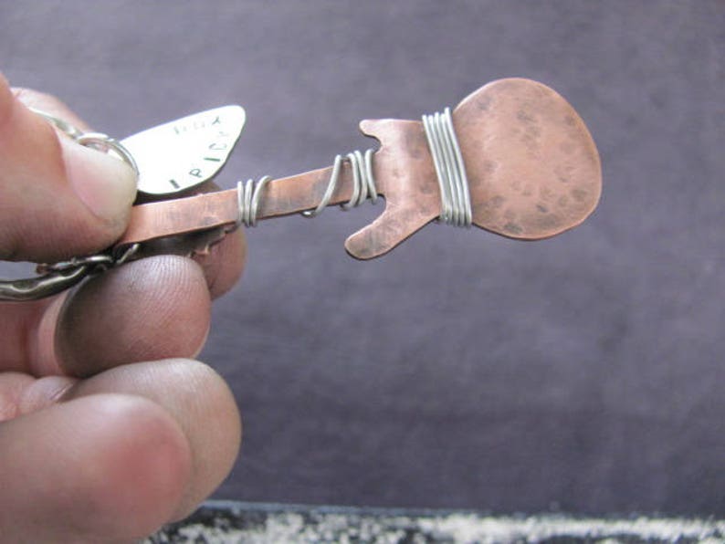 May include: A copper guitar-shaped pendant with a silver wire wrapped around the body. The pendant is attached to a silver pick with the word "I PICK" engraved on it.