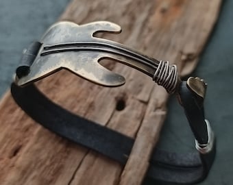May include: A bronze-colored guitar-shaped bracelet with a black leather band. The guitar has strings and is wrapped with silver wire at the ends. The bracelet is displayed on a piece of weathered wood.