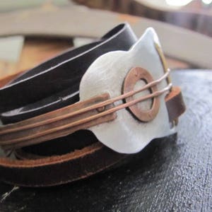 May include: A leather bracelet with a silver and copper guitar-shaped charm. The bracelet is made of three layers of leather, with the top layer being black, the middle layer being brown, and the bottom layer being a lighter brown. The guitar charm is attached to the bracelet with a copper band.