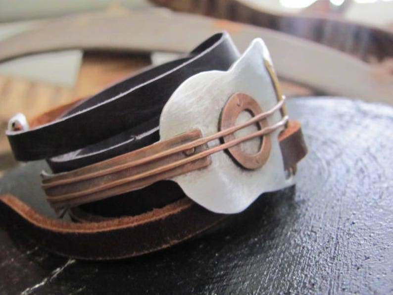 May include: A leather bracelet with a silver and copper guitar-shaped charm. The bracelet is made of three layers of leather, with the top layer being black, the middle layer being brown, and the bottom layer being a lighter brown. The guitar charm is attached to the bracelet with a copper band.