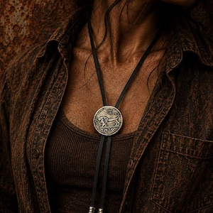 May include: A bolo tie featuring a circular silver-toned pendant with a lion relief and Hebrew text. The pendant is attached to a black leather cord with two silver-toned metal tips, each adorned with a small sun-shaped charm at the bottom.