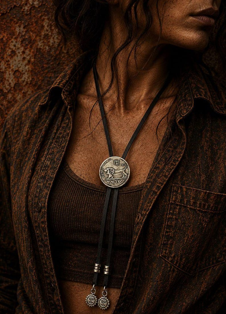May include: A bolo tie featuring a circular silver-toned pendant with a lion relief and Hebrew text. The pendant is attached to a black leather cord with two silver-toned metal tips, each adorned with a small sun-shaped charm at the bottom.