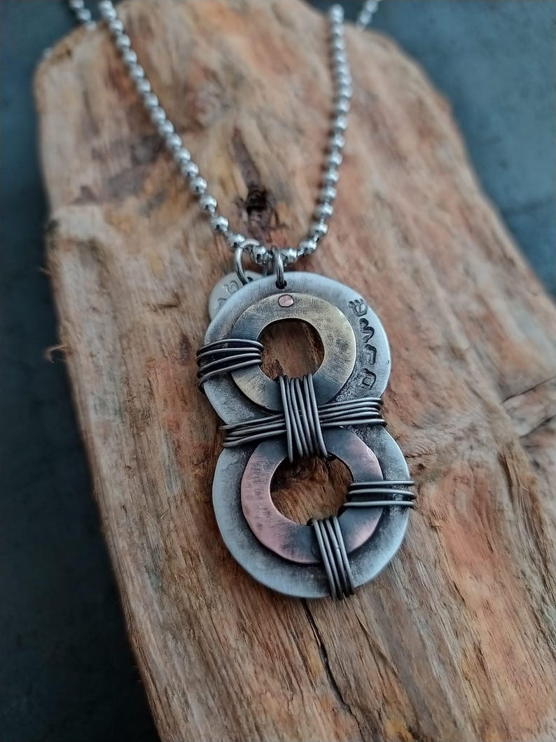 May include: A silver chain necklace with a pendant made of three metal rings. The rings are layered and connected with wire. The top ring is brass, the middle ring is copper, and the bottom ring is silver.