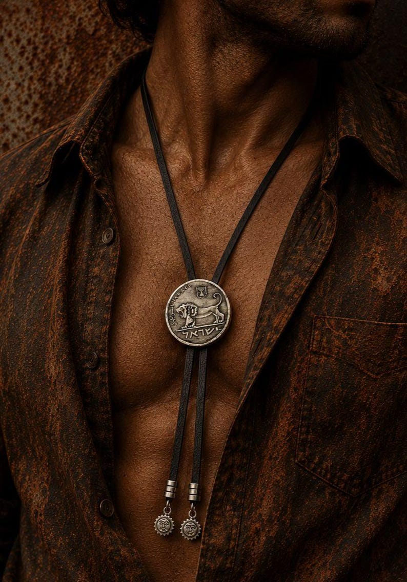 May include: A bolo tie featuring a circular silver-toned pendant with a relief of a lion and Hebrew text. The pendant is attached to a black leather cord that ends in two small silver-toned metal charms with sun-like designs at the bottom.