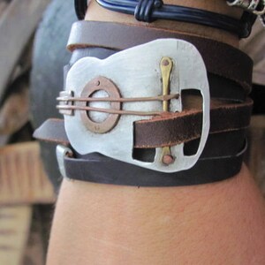 May include: A leather bracelet with a silver guitar-shaped buckle. The buckle has a copper-colored accent and a small hole in the center.