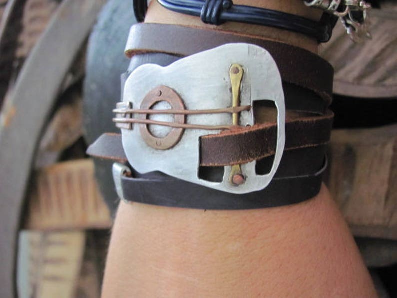 May include: A leather bracelet with a silver guitar-shaped buckle. The buckle has a copper-colored accent and a small hole in the center.