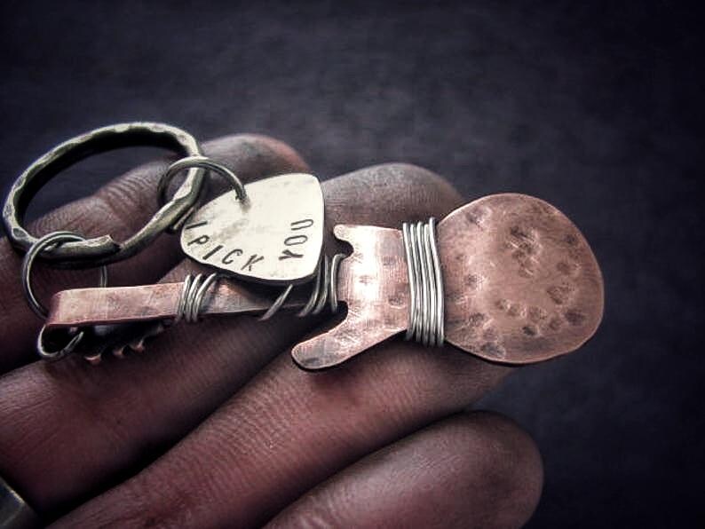 May include: A silver guitar pick keychain with the words "I pick you" engraved on it. The keychain is attached to a copper guitar-shaped charm with a hammered finish.