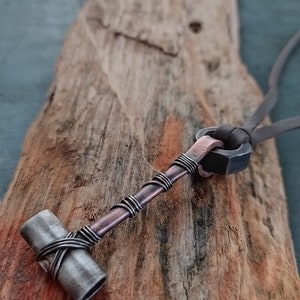 May include: A copper and silver necklace with a hammer pendant. The hammer is made of metal and has a copper wire wrapped around it. The necklace is on a wooden surface.