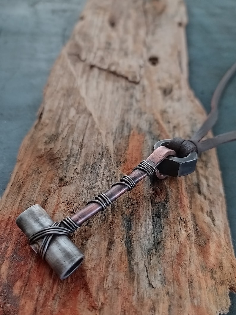 May include: A copper and silver necklace with a hammer pendant. The hammer is made of metal and has a copper wire wrapped around it. The necklace is on a wooden surface.