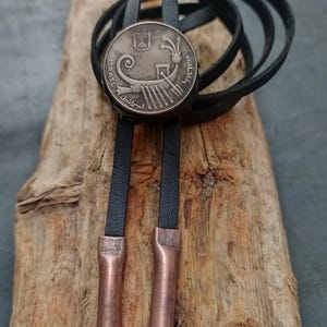 May include: A black leather cord bracelet with a silver coin charm featuring a ship design and the text "ISRAEL" and "ישראל". The cord ends have copper tips.
