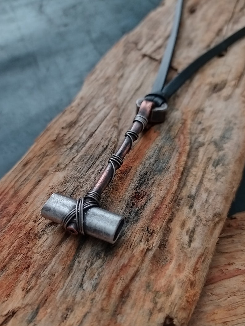 May include: A silver and copper wire wrapped pendant in the shape of a hammer, hanging from a black leather cord.