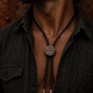 May include: A bolo tie featuring a circular, weathered bronze-toned medallion with an engraved scorpion design. The medallion is centered on a thin black cord that hangs down, finished with two matching metallic tips at the bottom ends of the cord.