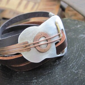 May include: A leather bracelet with a silver guitar-shaped charm. The charm has a copper band around it and a copper circle in the center.