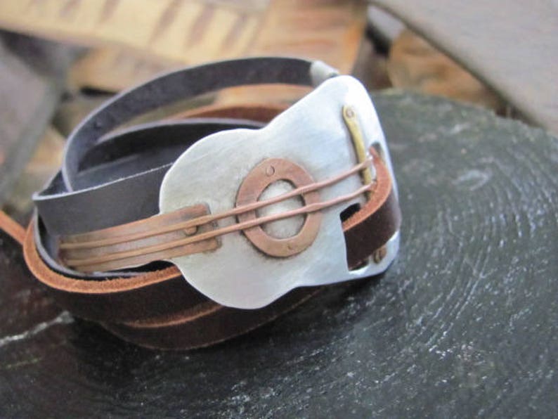 May include: A leather bracelet with a silver guitar-shaped charm. The charm has a copper band around it and a copper circle in the center.