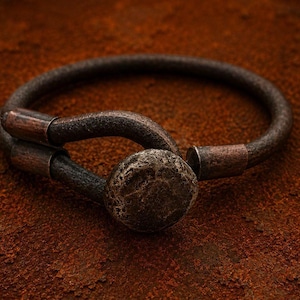 May include: A dark brown, textured circular bracelet featuring a hook-and-eye style closure. The bracelet has a weathered, metallic appearance with copper-toned accents at the ends and a central, rounded, rough-surfaced clasp mechanism.