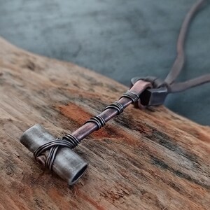 May include: A silver and copper necklace with a hammer pendant. The hammer is made of metal and has a copper wire wrapped around it. The necklace is on a wooden surface.