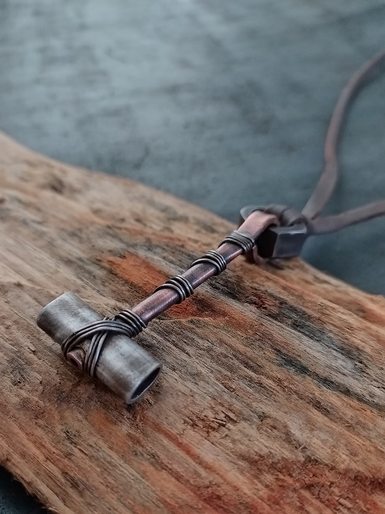 May include: A silver and copper necklace with a hammer pendant. The hammer is made of metal and has a copper wire wrapped around it. The necklace is on a wooden surface.
