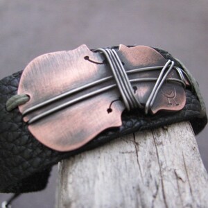May include: A black leather bracelet with a copper and silver violin-shaped charm. The charm is decorated with wire wrapped around it.