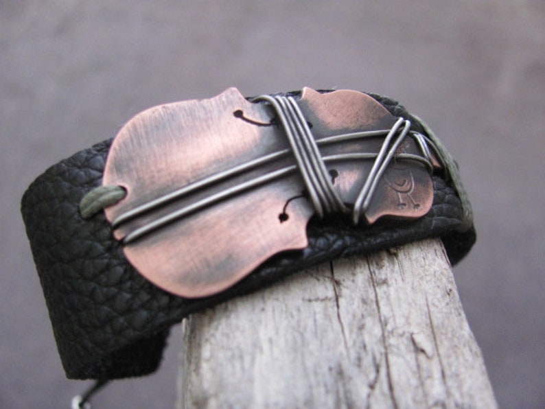 May include: A black leather bracelet with a copper and silver violin-shaped charm. The charm is decorated with wire wrapped around it.