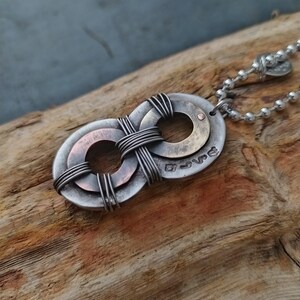 May include: A silver infinity symbol necklace with a copper and brass accent. The pendant is made of two intertwined circles with a wire wrapped around them. The necklace is on a silver chain.