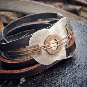 May include: A leather bracelet with a silver and copper guitar-shaped charm. The charm has a circular design with a copper center and a silver outer ring.