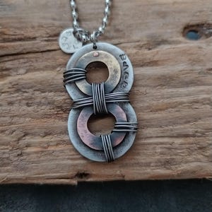 May include: A silver chain necklace with a pendant made of two intertwined metal circles. The circles are layered with copper and silver, and the top circle has the Hebrew word "שלום" inscribed on it.