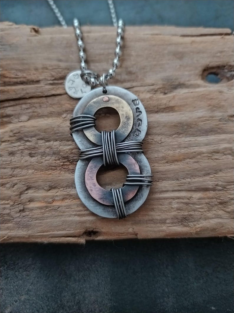 May include: A silver chain necklace with a pendant made of two intertwined metal circles. The circles are layered with copper and silver, and the top circle has the Hebrew word "שלום" inscribed on it.