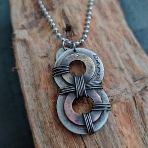 May include: A silver chain necklace with a pendant made of three metal rings. The rings are layered and connected with wire. The top ring is brass, the middle ring is copper, and the bottom ring is silver.