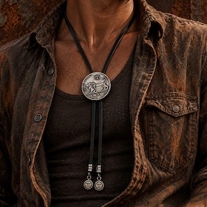 May include: A bolo tie featuring a circular silver-toned pendant with an embossed lion and Hebrew text. The pendant is attached to a black leather cord that ends in two silver-toned metal tips, each decorated with a small sun-like face emblem.