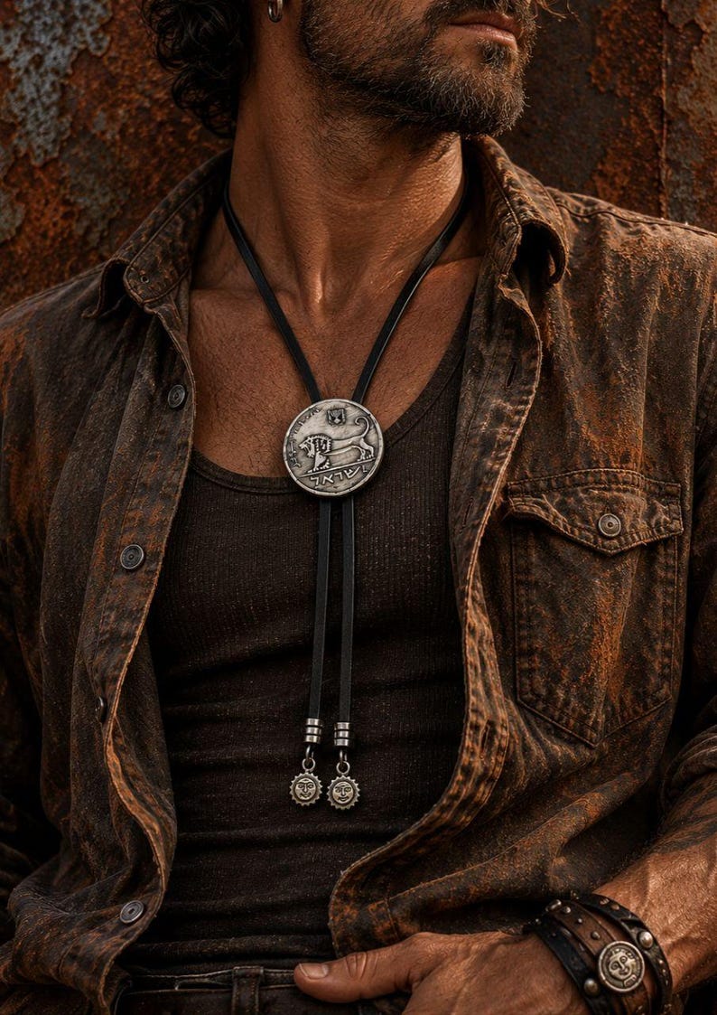 May include: A bolo tie featuring a circular silver-toned pendant with an embossed lion and Hebrew text. The pendant is attached to a black leather cord that ends in two silver-toned metal tips, each decorated with a small sun-like face emblem.
