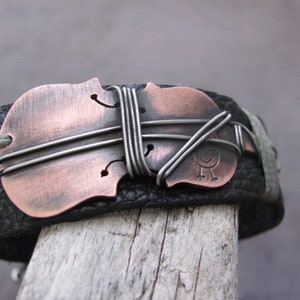 May include: A black leather bracelet with a copper-colored violin charm. The charm is decorated with silver wire and a small bird design.