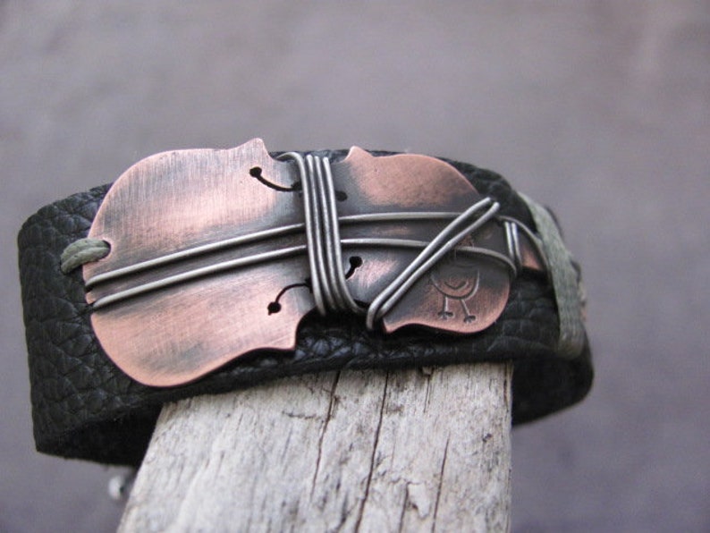 May include: A black leather bracelet with a copper-colored violin charm. The charm is decorated with silver wire and a small bird design.