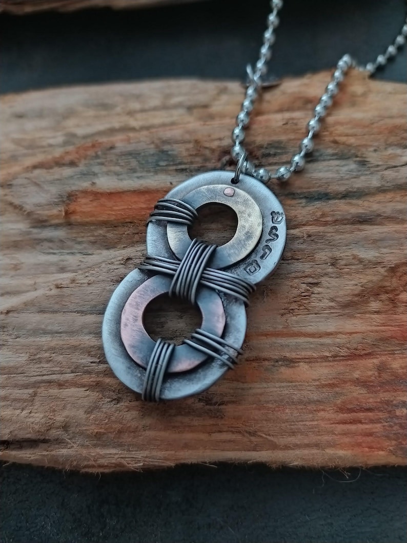 May include: A silver and copper necklace with a double ring pendant. The rings are intertwined with wire and have a small copper dot in the center. The necklace is on a wooden surface.