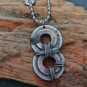 May include: A silver chain necklace with a pendant made of two intertwined metal rings. The rings are layered, with the top ring being copper and the bottom ring being silver. The pendant has a small dot of copper in the center.