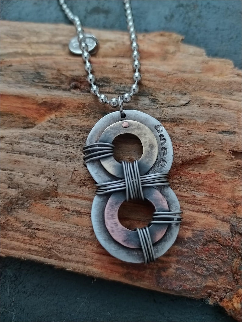 May include: A silver chain necklace with a pendant made of two intertwined metal rings. The rings are layered, with the top ring being copper and the bottom ring being silver. The pendant has a small dot of copper in the center.