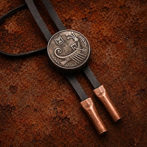 May include: A bolo tie featuring a circular silver-toned metal pendant with an embossed ancient lyre design, the word ISRAEL, and a menorah emblem. The pendant is attached to a black leather cord finished with two cylindrical copper-toned metal tips.