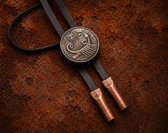 Könnte beinhalten: Eine Bolo-Krawatte mit einem runden, silberfarbenen Metallanhänger, der ein geprägtes antikes Leier-Design, das Wort ISRAEL und ein Menora-Emblem zeigt. Der Anhänger ist an einer schwarzen Lederschnur mit zwei zylindrischen, kupferfarbenen Metallenden befestigt.