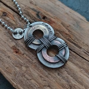 May include: A silver chain necklace with a pendant made of two overlapping metal circles. The circles are wrapped with wire and have a copper and brass finish. The pendant is on a wooden surface.