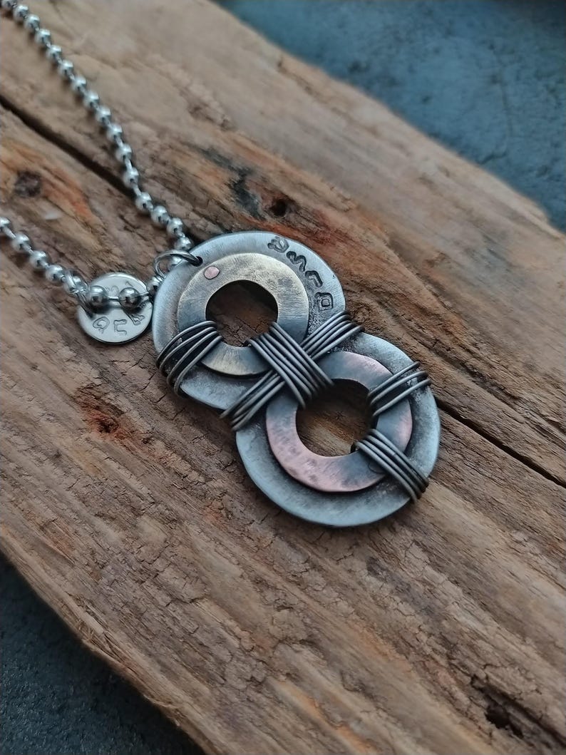May include: A silver chain necklace with a pendant made of two overlapping metal circles. The circles are wrapped with wire and have a copper and brass finish. The pendant is on a wooden surface.