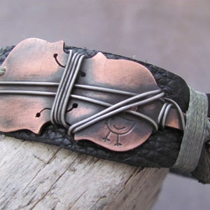 May include: A black leather bracelet with a copper-colored violin design and wire accents. The violin is wrapped with silver wire and has a small bird design on the body.