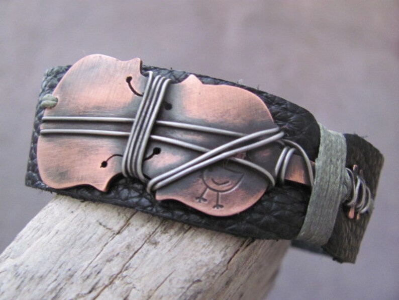 May include: A black leather bracelet with a copper-colored violin design and wire accents. The violin is wrapped with silver wire and has a small bird design on the body.