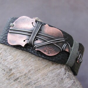 May include: A black leather bracelet with a copper-colored violin charm. The charm is wrapped with silver wire and has a small bird design.