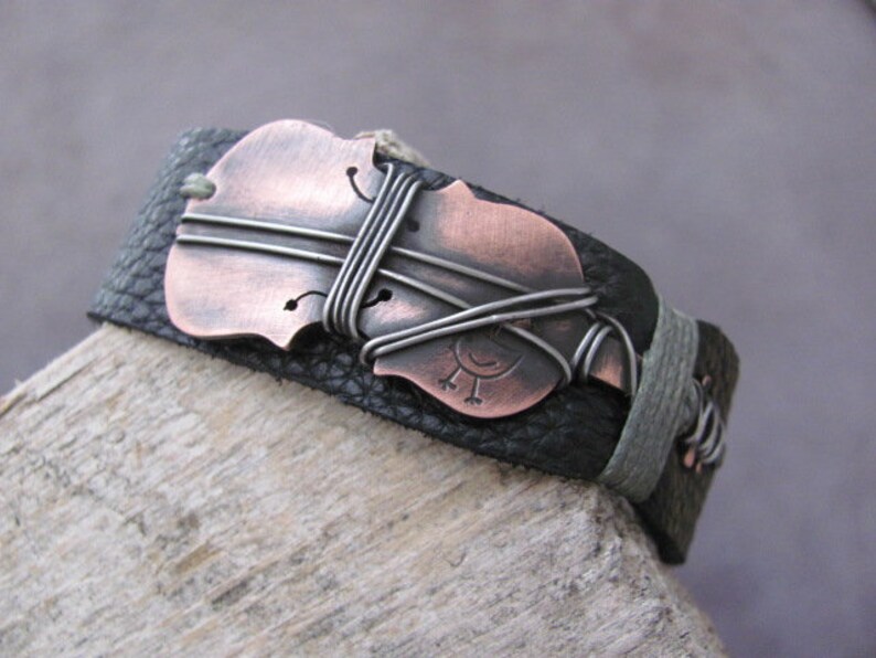 May include: A black leather bracelet with a copper-colored violin charm. The charm is wrapped with silver wire and has a small bird design.