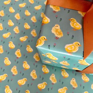 Peut inclure: Papier cadeau avec un motif r&eacute;p&eacute;t&eacute; de poussins jaunes sur fond bleu sarcelle. Une bo&icirc;te cadeau emball&eacute;e avec un motif assorti est nou&eacute;e avec un large ruban orange. Le motif comprend de petits accents floraux.