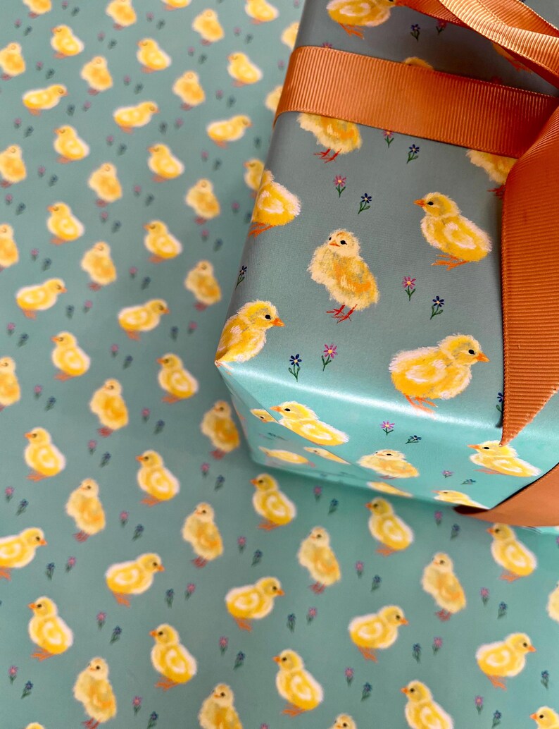 Peut inclure: Papier cadeau avec un motif r&eacute;p&eacute;t&eacute; de poussins jaunes sur fond bleu sarcelle. Une bo&icirc;te cadeau emball&eacute;e avec un motif assorti est nou&eacute;e avec un large ruban orange. Le motif comprend de petits accents floraux.