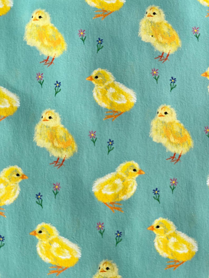 Peut inclure: Motif r&eacute;p&eacute;t&eacute; de poussins jaunes avec des becs et des pattes orange sur fond bleu clair. De petites fleurs violettes et bleues sont dispers&eacute;es dans le motif. Les poussins sont peints avec un effet de coups de pinceau textur&eacute;s.